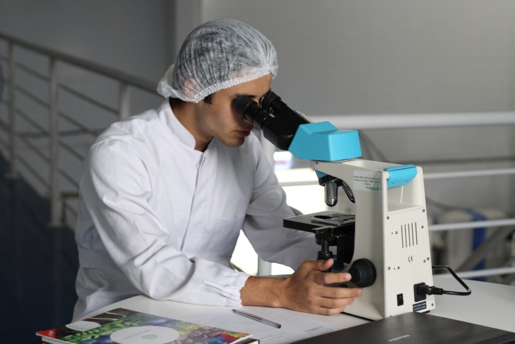 Man wearing a lab coat and hair net looking into a microscope.