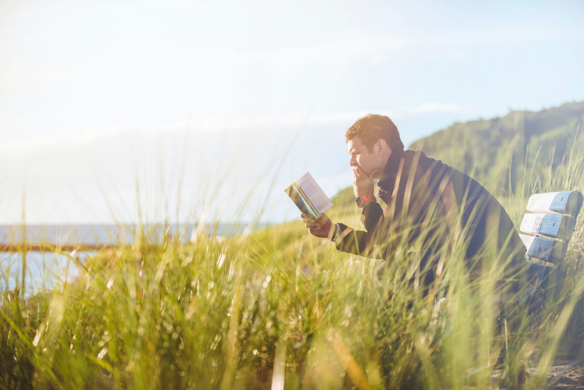 Man reading a book while sitting on a bench in a park near a lake.