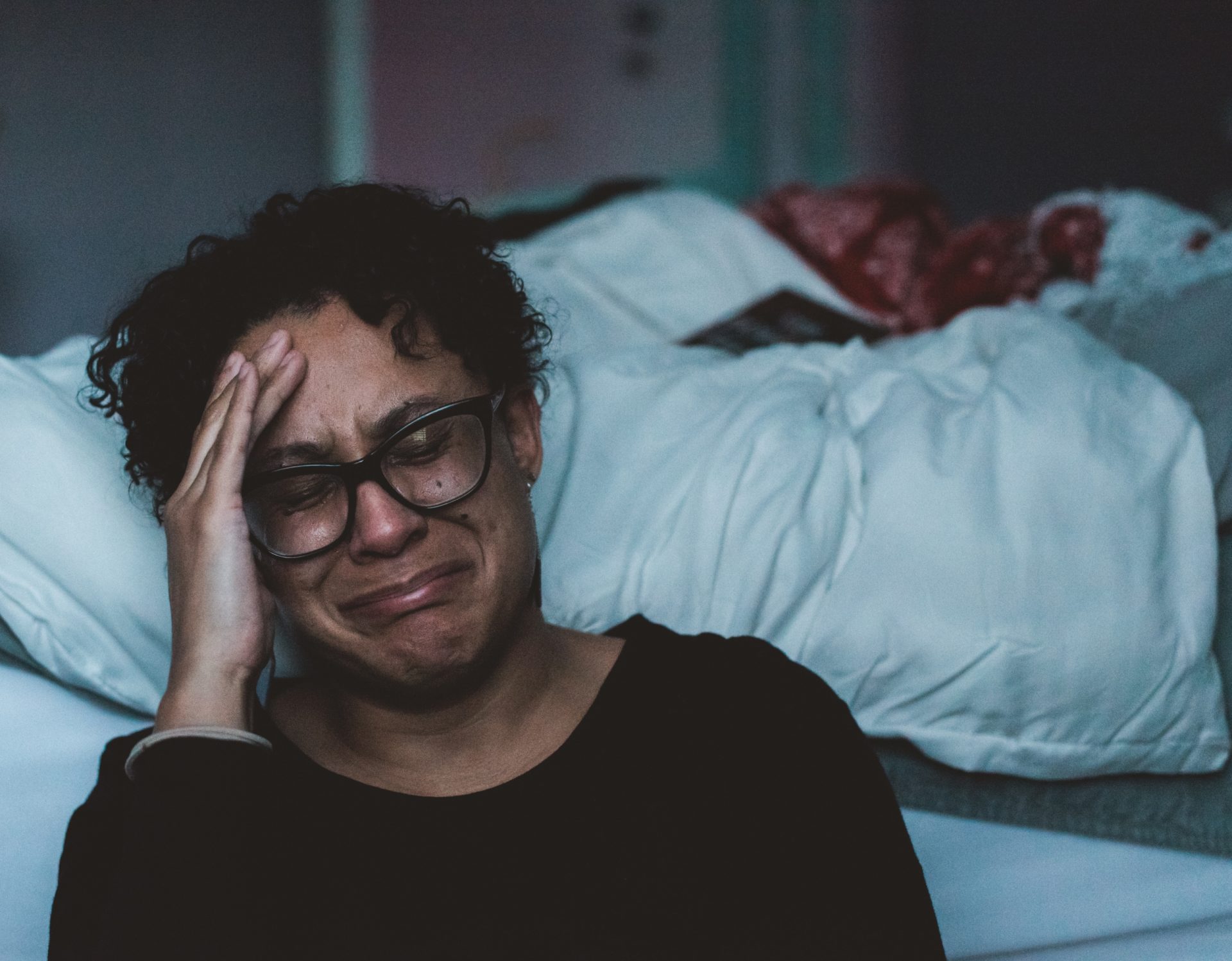 Woman sitting on the floor, leaning against her unmade bed and crying.