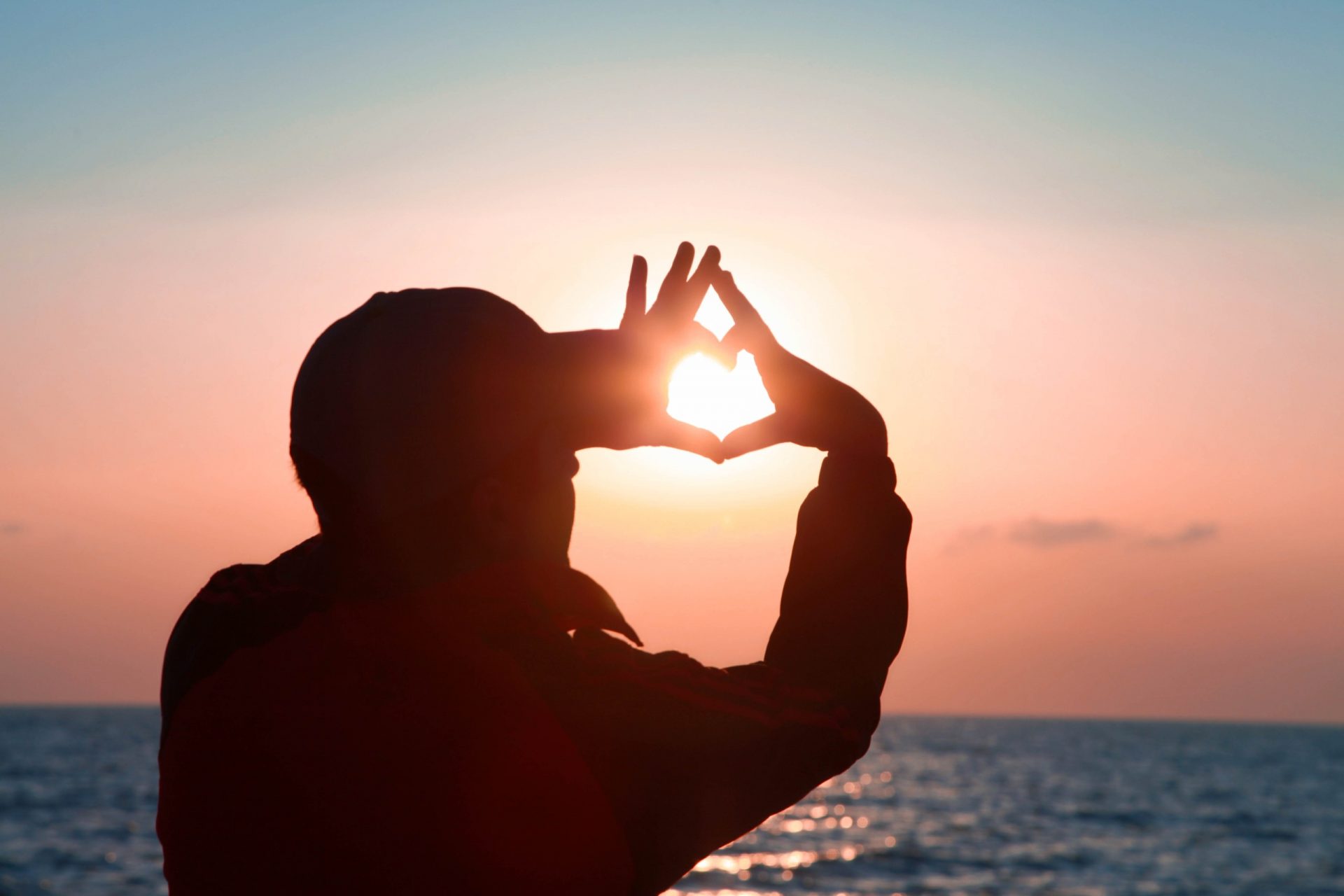 Man holding his hands in the shape of a heart over the setting sun.