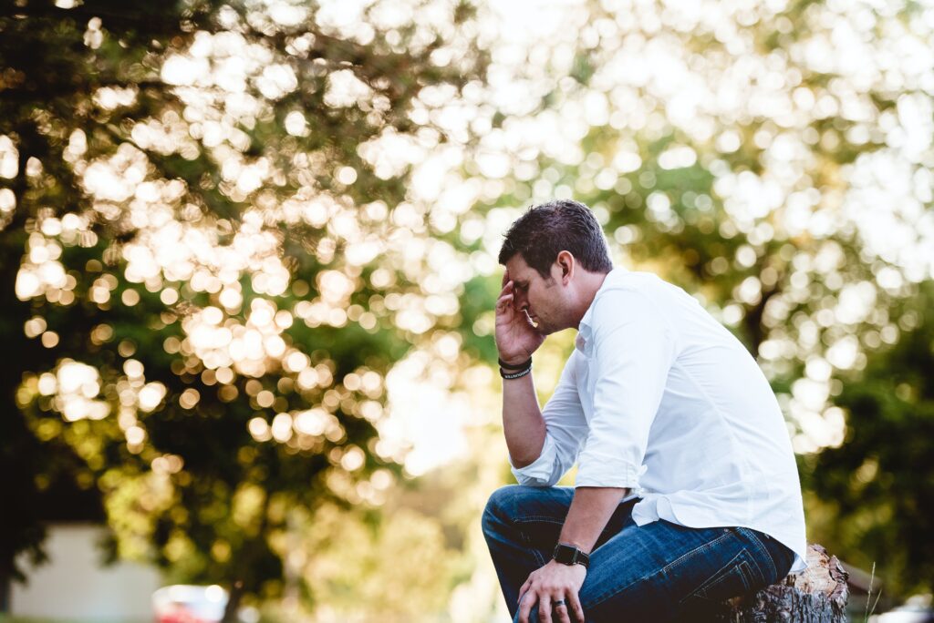 Man sitting on a stump holding his head as he struggles with anxiety, aches and pains everywhere.