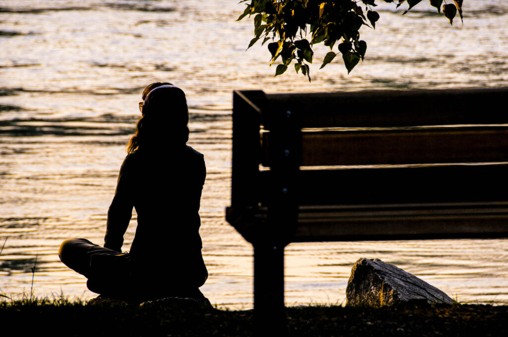Woman sitting cross-legged on a rock and looking out at placid lake at sunset.