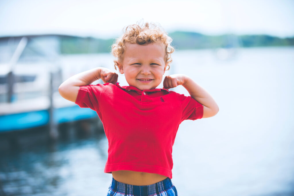 Little boy standing on a dock and showing off his muscles.