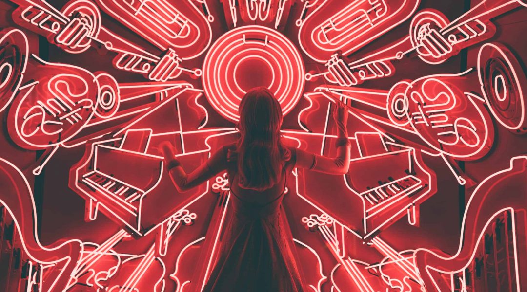 Woman standing in front of neon images of musical instruments pretending to conduct them.