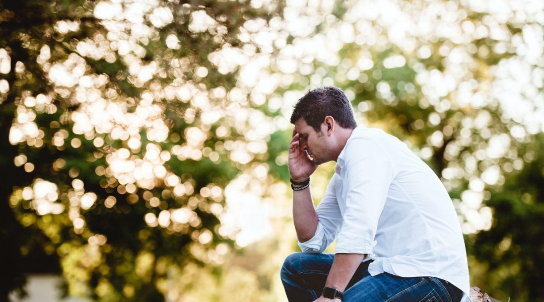 Man sitting on a stump holding his head as he struggles with anxiety, aches and pains everywhere.