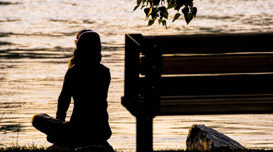Woman sitting cross-legged on a rock and looking out at placid lake at sunset.