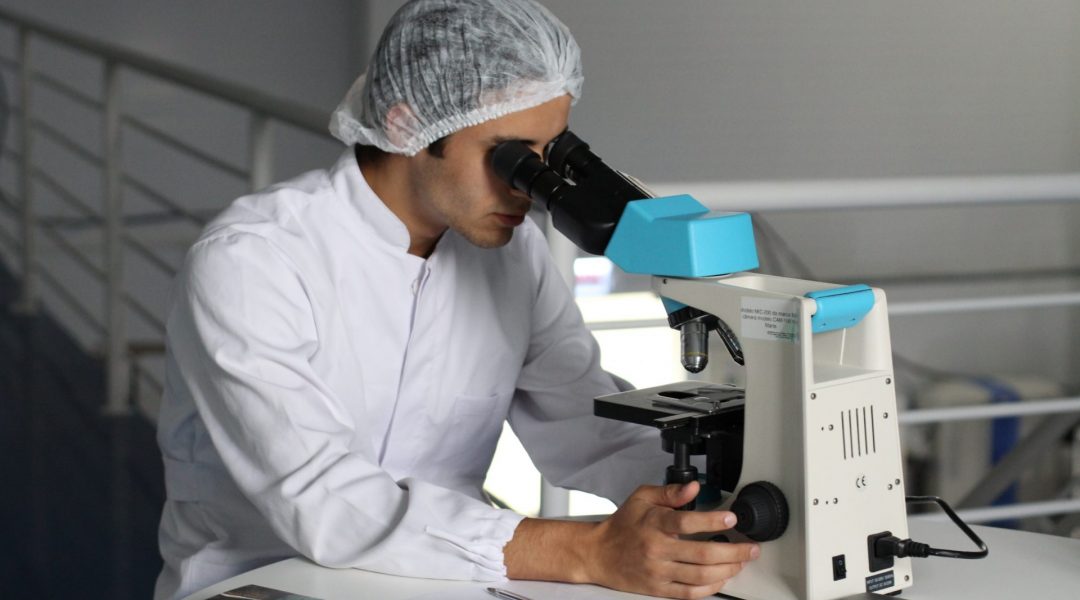 Man wearing a lab coat and hair net looking into a microscope.