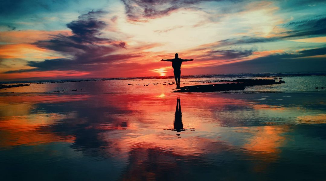 Man standing on the shore with his arms outstretched as he watches the sunrise.